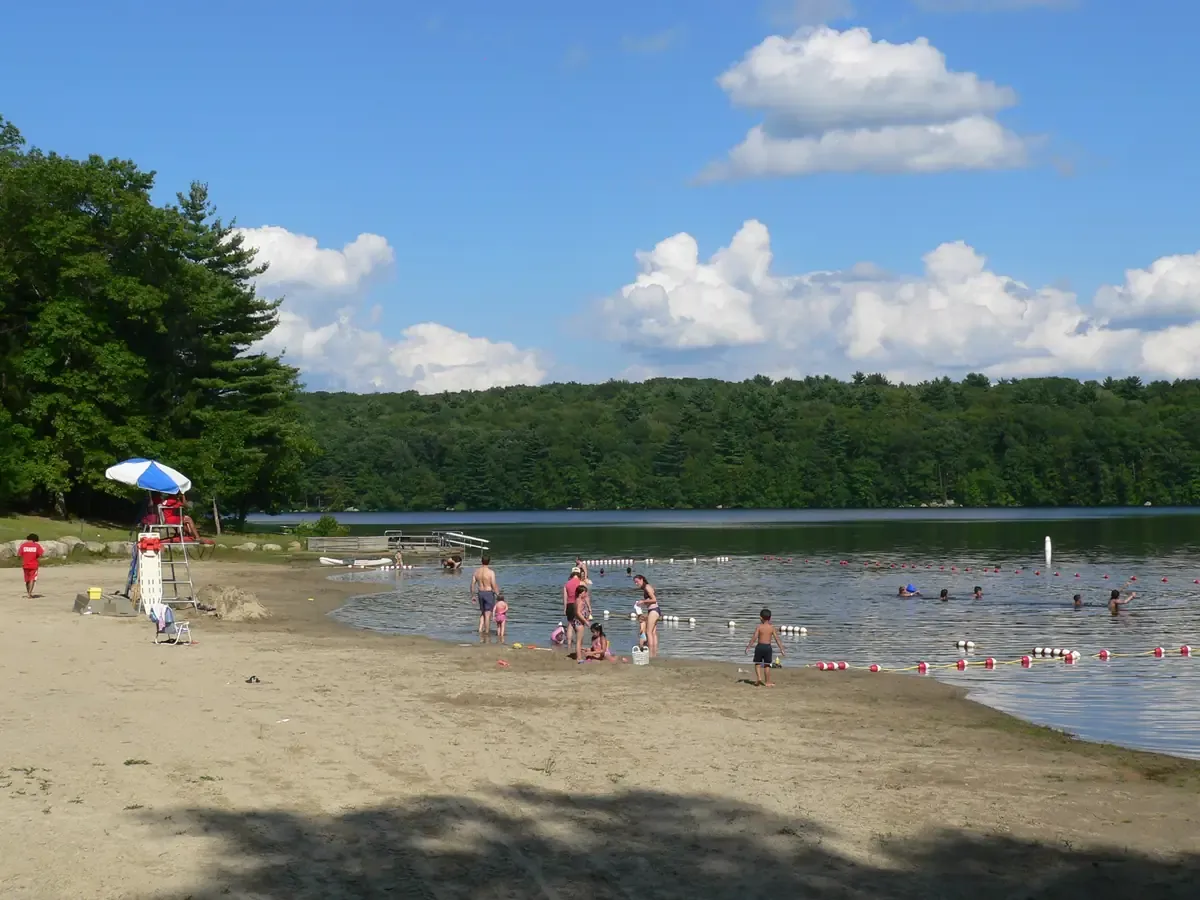 Families on the sandy swimming beach at Ashland State Park, with the reservoir and treeline behind