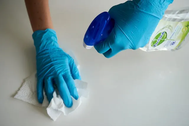 Gloved hands wiping a white surface with a cloth and a spray bottle