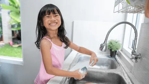 A child smiling while washing a dish at the kitchen sink